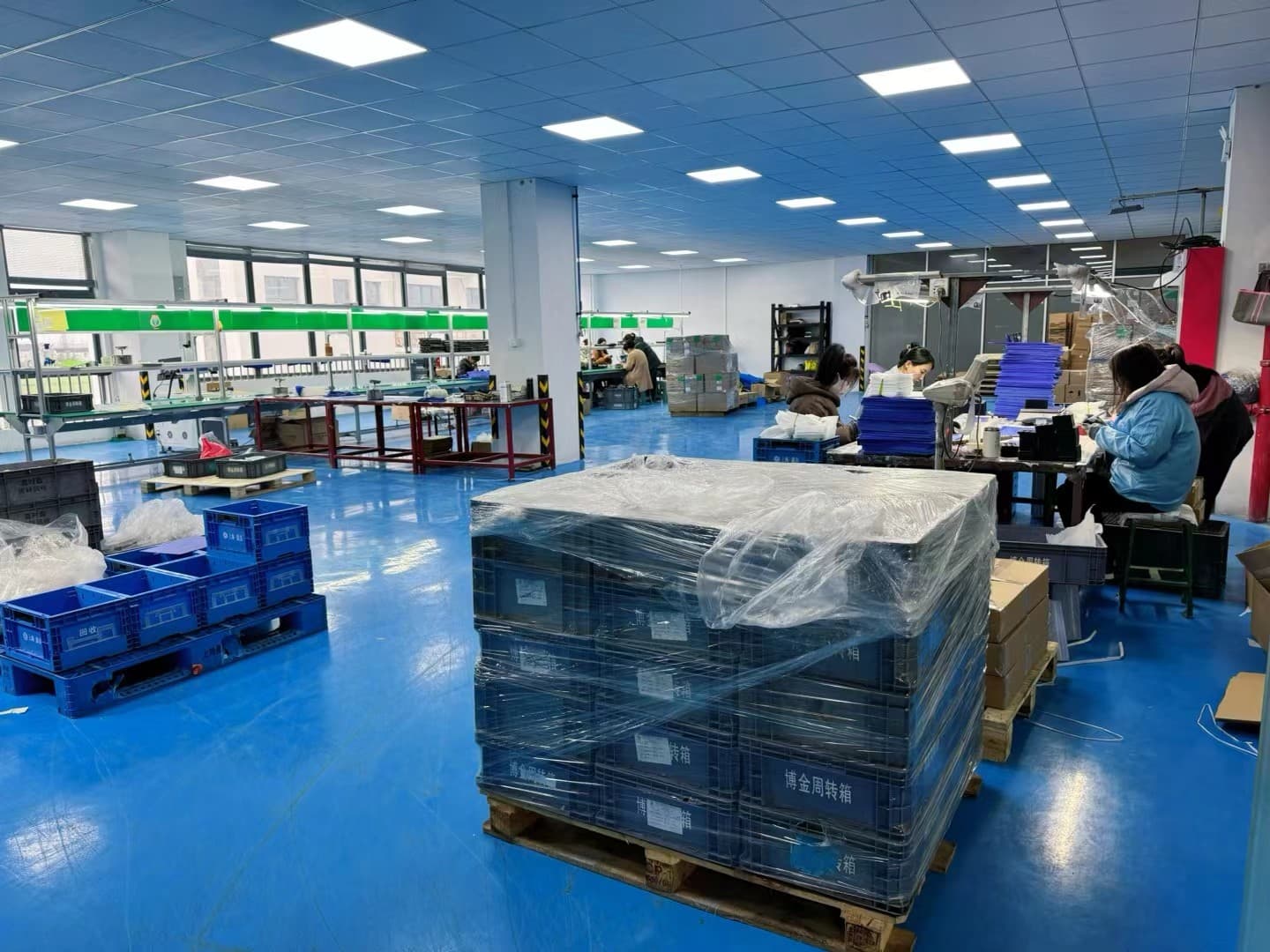 Assembly benches with operators and a wrapped pallet of blue turnover boxes in the packing area